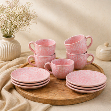 Pink ceramic tea set with six cups and matching saucers arranged on a round wooden tray, placed on a neutral beige background with a vase and soft fabric for a minimal, elegant setup.